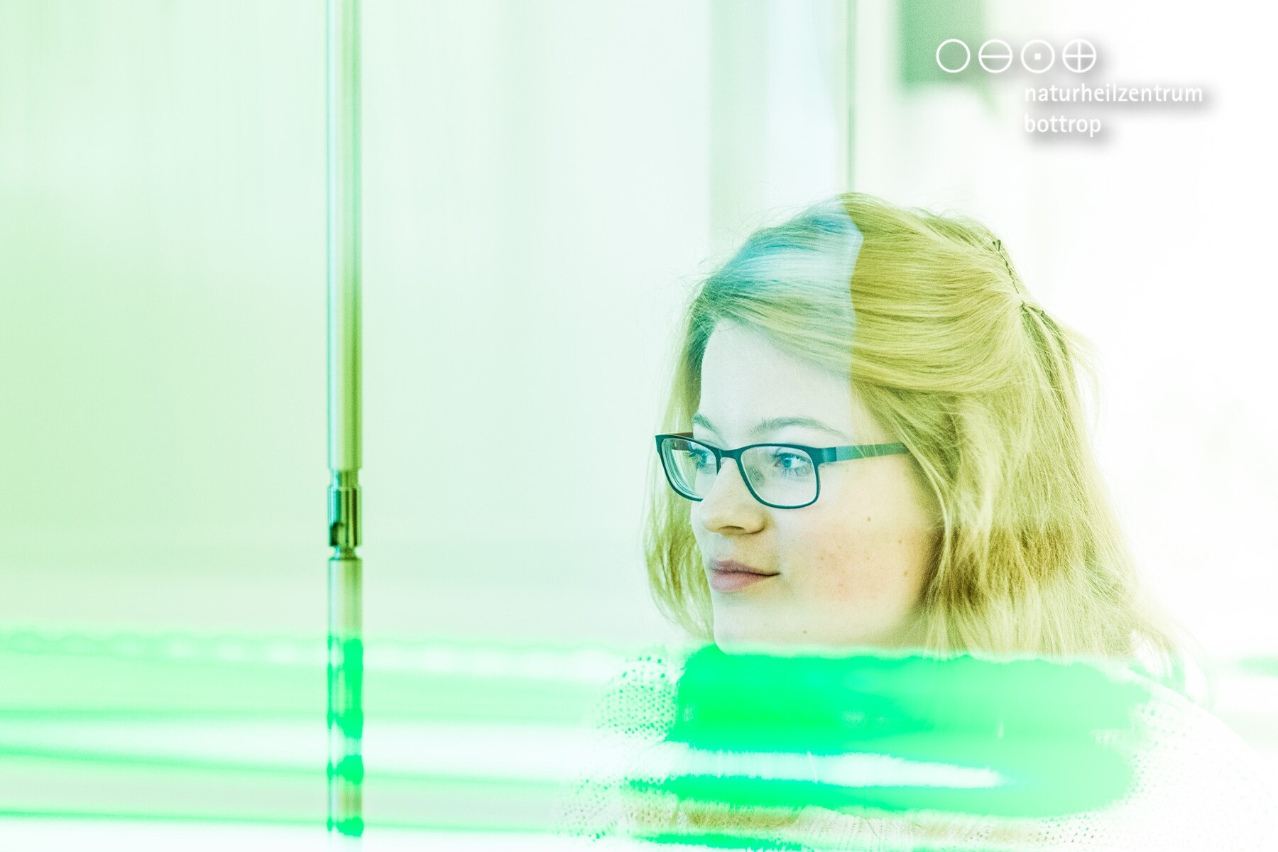 Eine Person mit blonden, lockigen Haaren und Brille, die nachdenklich aussieht, vor einem grünen Licht und reflektierenden Oberflächen.