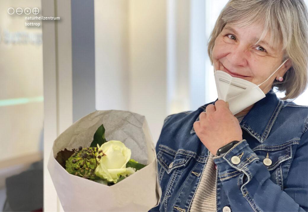 Woman in denim jacket with FFP2 mask pulled down presents a bouquet of flowers.