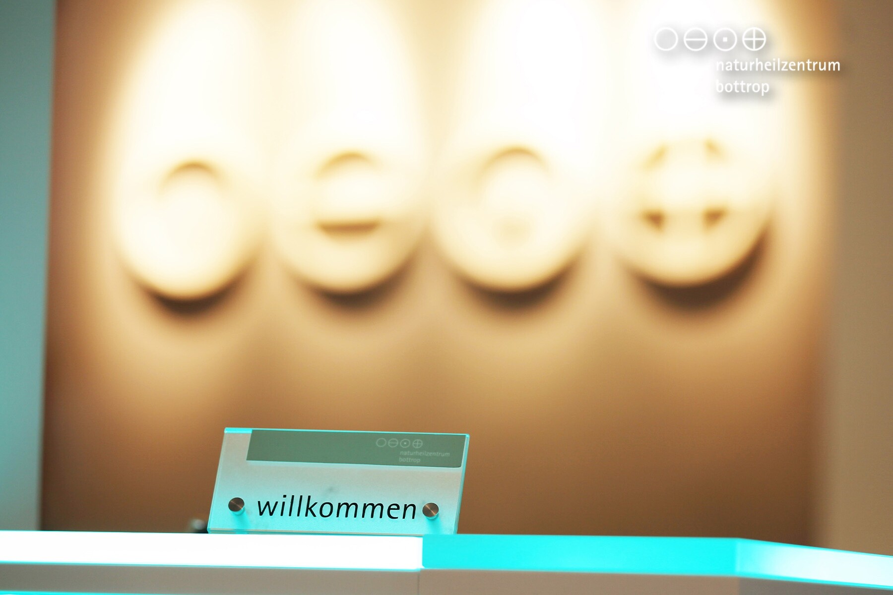 Ein Empfangstresen mit einem Schild, auf dem willkommen steht, vor einer unscharfen Wand mit symbolischen Abbildungen und dem Logo des Naturheilzentrums Bottrop im Hintergrund.